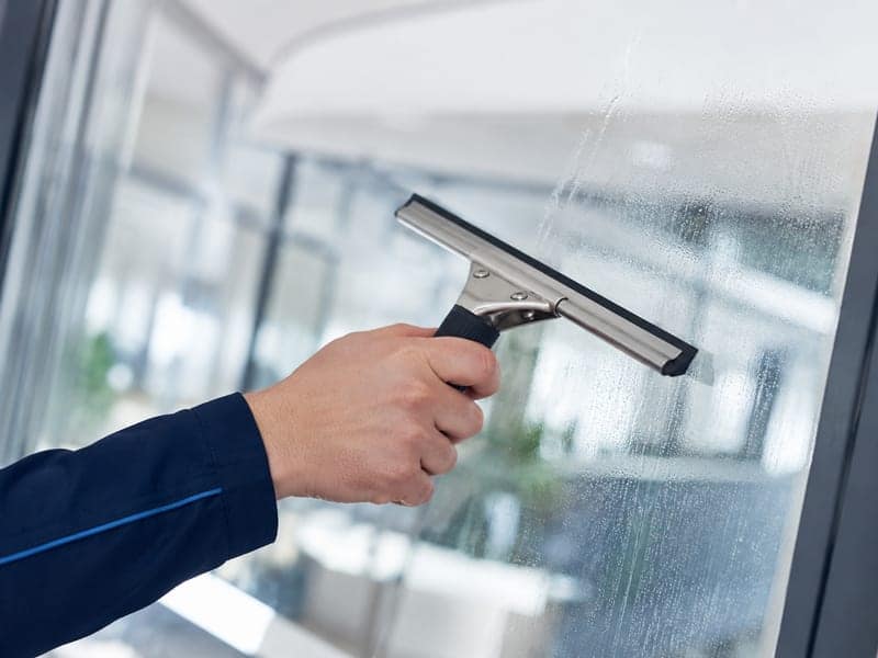 Hand mit professionellem Fensterabzieher zieht eine Bahn über eine Glasscheibe, der abgezogene Streifen ist klar